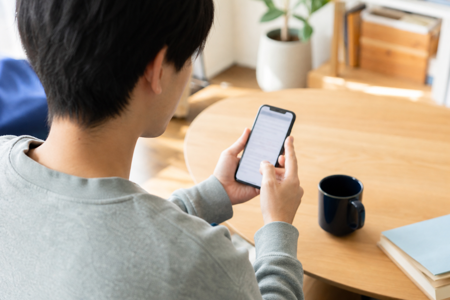 単身者のスマホ相談：名古屋・愛知で学生（親が連帯保証人になれない／実家が遠方で印鑑証明を出せない）が賃貸申込をする際、審査を通すために準備すべき書類・順序・不動産会社への伝え方を知りたいについて一人でスマートフォン確認をする場面を斜め後方から撮る。画面はぼかし、手元中心、カップルなし